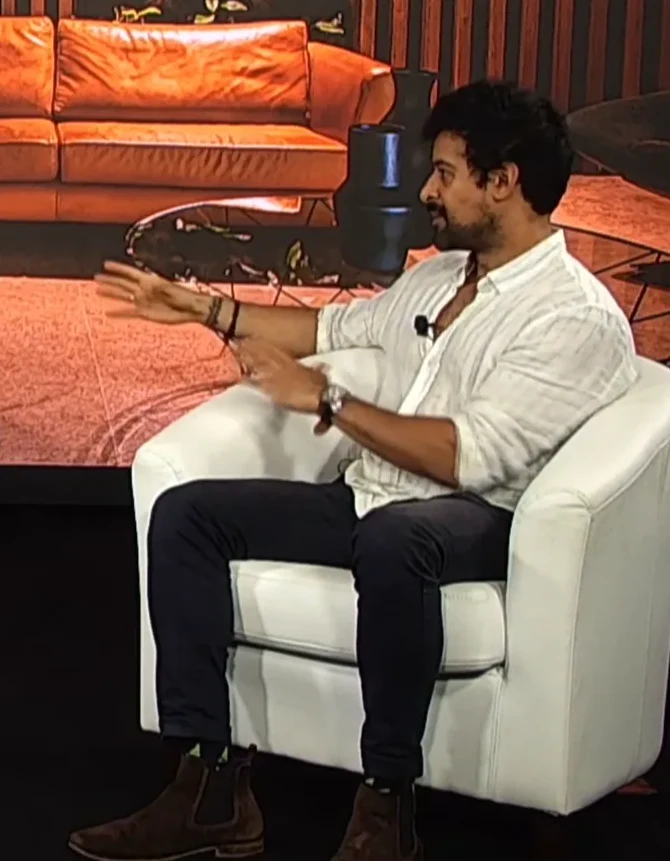 Krish Raja on stage, seated and mid-sentence with a hand raised.